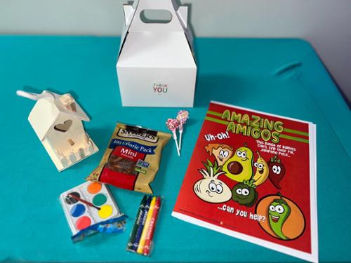 a white box with "thank you" sticker, include wooden house, watercolors, coloring book, crayons, pretzel, two lollypops.